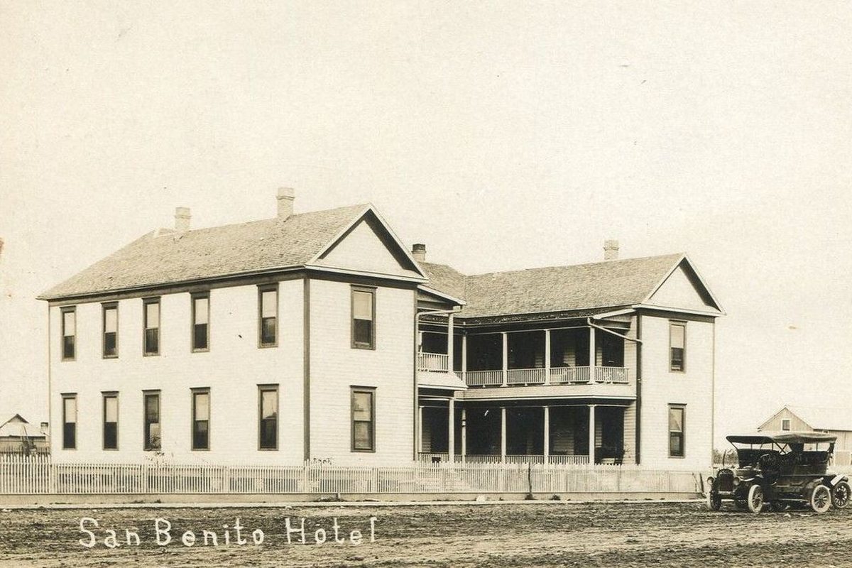 Old sepia postcard of the two-story San Benito Hotel with long porches and a picket fence, an early open-top automobile parked on the dirt street beside it