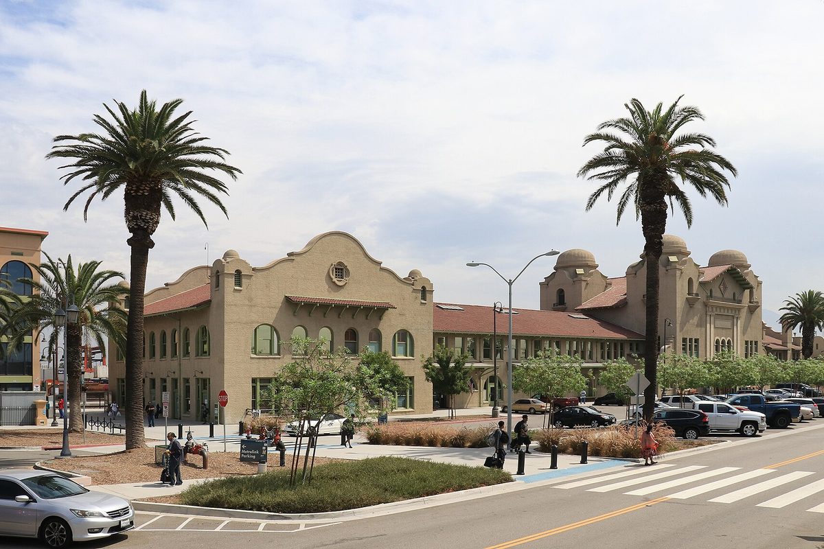 The cream-colored Mission Revival Santa Fe Depot in San Bernardino, with arched windows and domed towers, behind tall palm trees, parked cars and a crosswalk