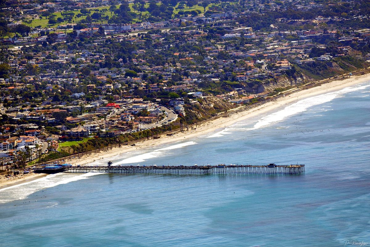 Aerial view of the San Clemente Pier reaching into blue Pacific water, with the beach and hillside neighborhoods behind it