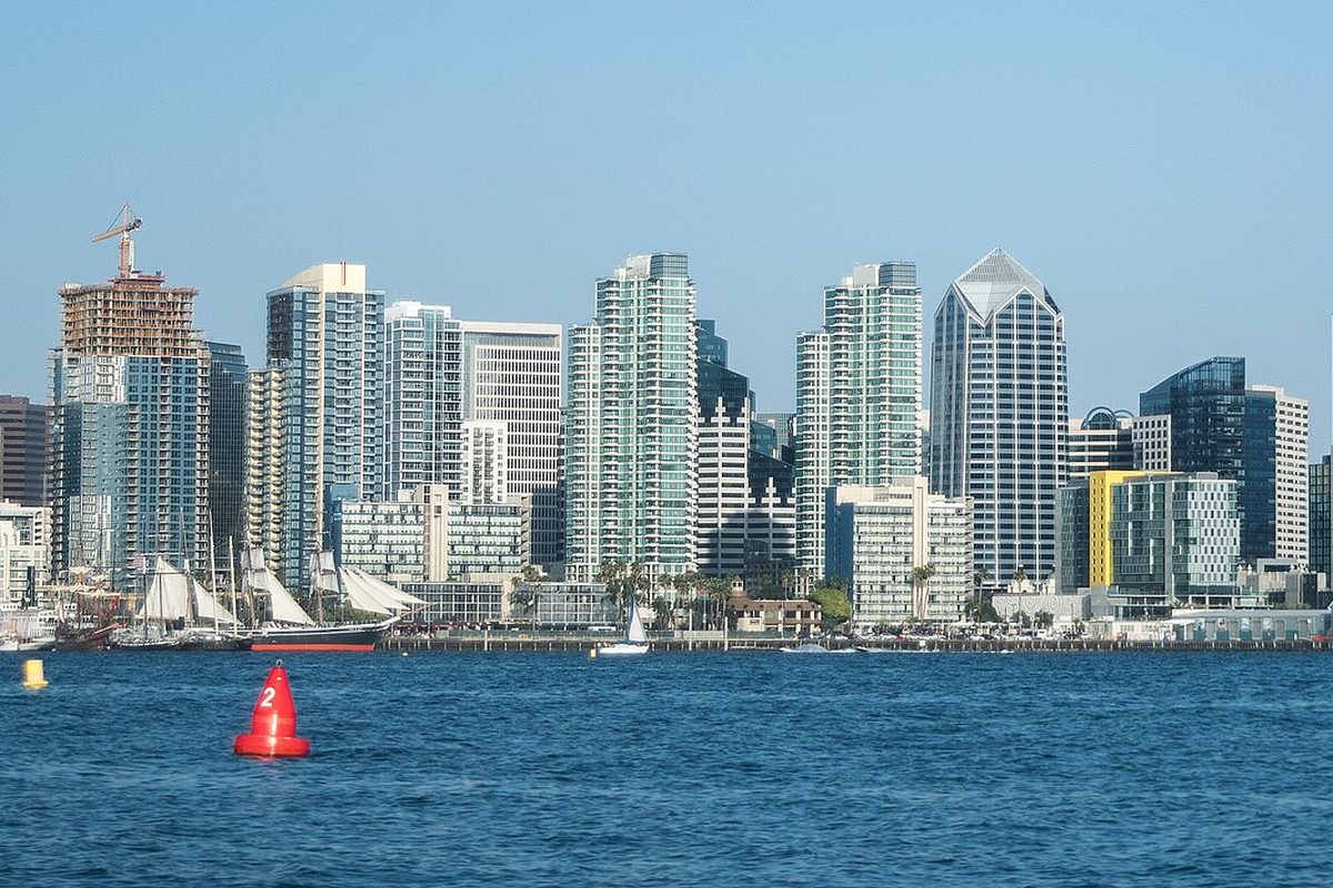 Downtown San Diego high-rises seen across the blue water of San Diego Bay, with a red channel buoy in front and a tall ship's white sails at the waterfront