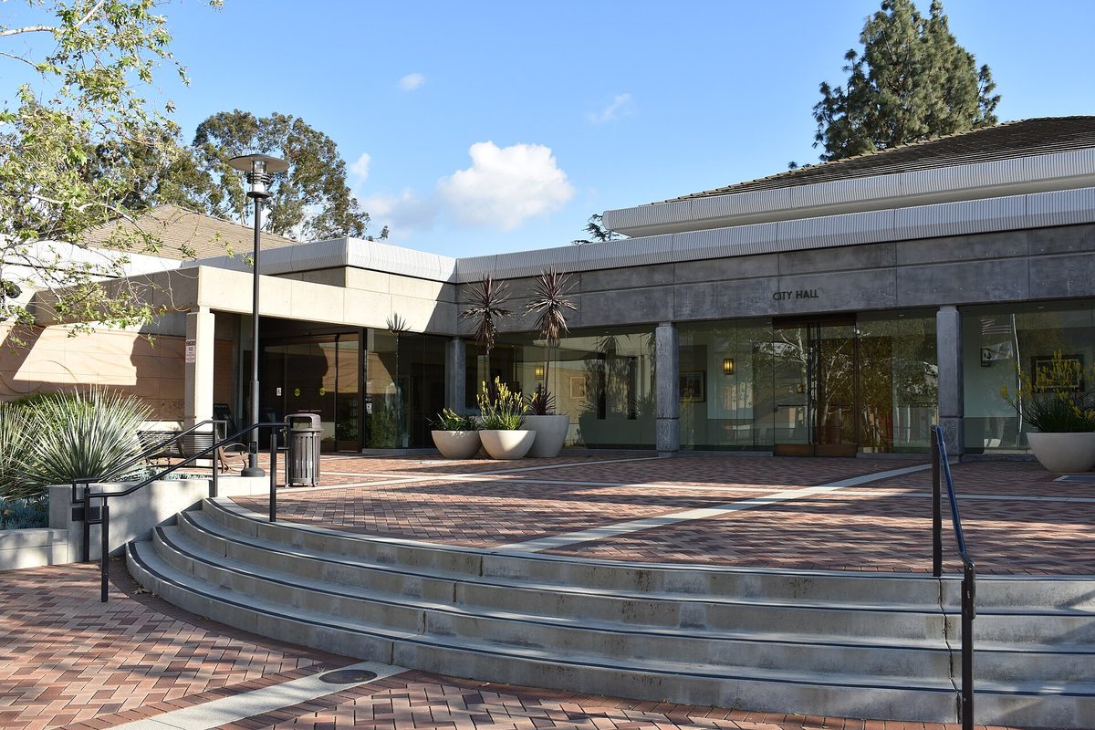 San Dimas City Hall, a low building with a glass front and a CITY HALL sign, above a brick plaza with curved concrete steps, potted plants and pine trees