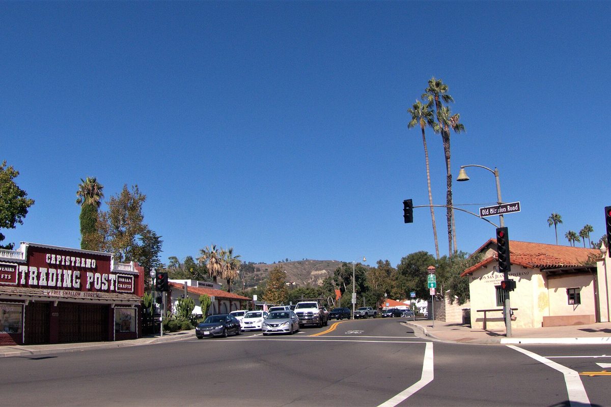 The Capistrano Trading Post storefront on Camino Capistrano at the Old Mission Road signal, with tall palms, parked cars and brown hills under a clear blue sky