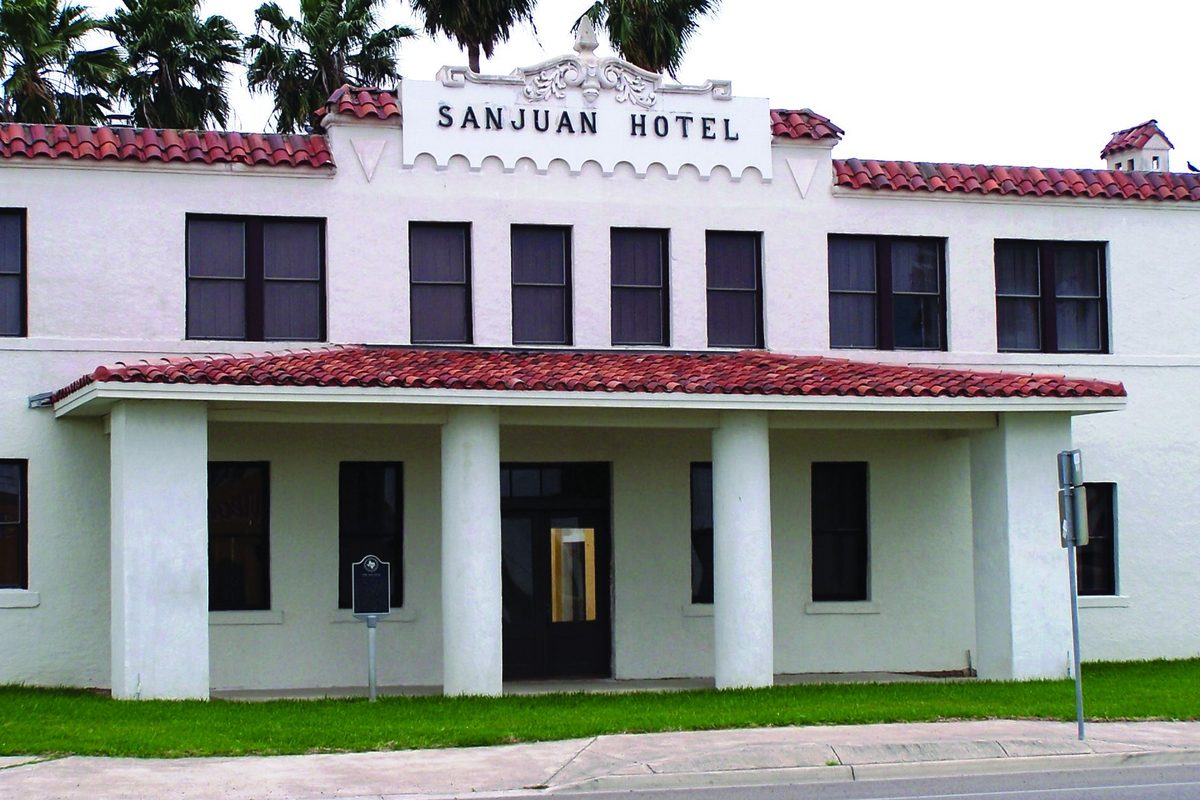 The white two-story San Juan Hotel with a red tile roof, square columns along a covered porch, a Texas historical marker on the lawn and palm trees behind