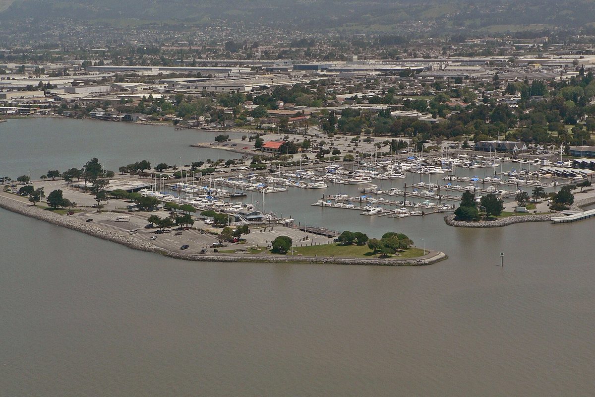 Aerial view of the San Leandro Marina, rows of boats inside a curved rock breakwater on the bay, with the town spreading inland behind it