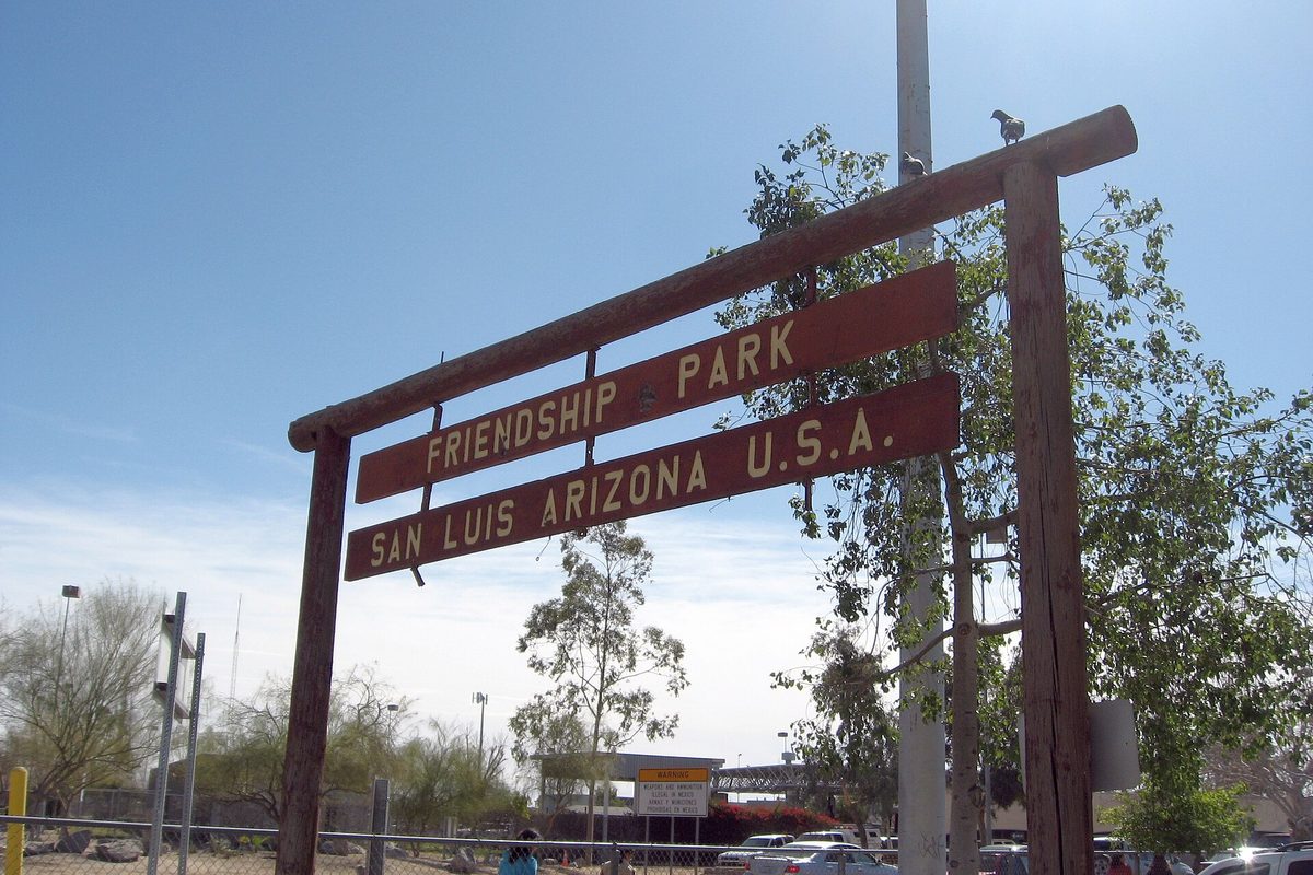 A wooden sign reading Friendship Park, San Luis Arizona U.S.A. under a blue sky, with trees and a parking area behind it