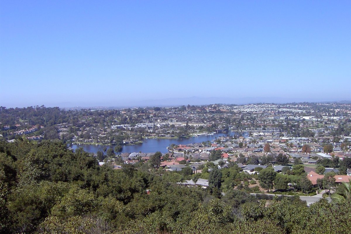 Hillside view over San Marcos, with neighborhoods of homes spread around a blue lake under a clear sky and green brush in the foreground