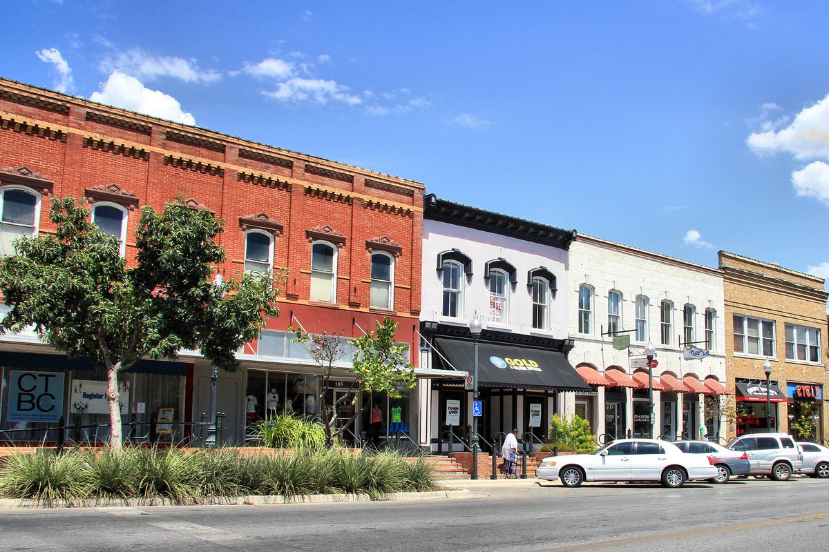 Red brick and white two-story storefronts along East Hopkins Street in downtown San Marcos, with ornamental grasses in the median and cars parked at the curb