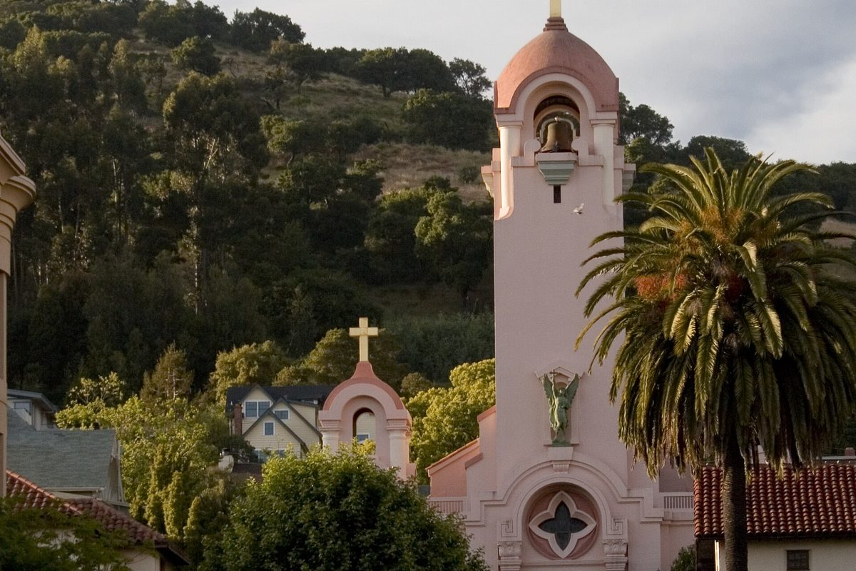 The pink bell tower of Saint Raphael Church in San Rafael beside a tall palm tree, with a wooded hillside behind it