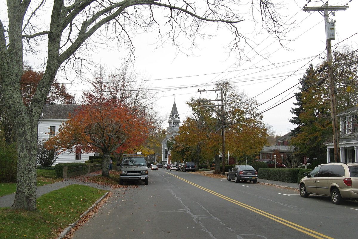 Main Street in Sandwich in autumn, orange-leaved trees and a white church steeple ahead, a pickup and a minivan parked along the curb