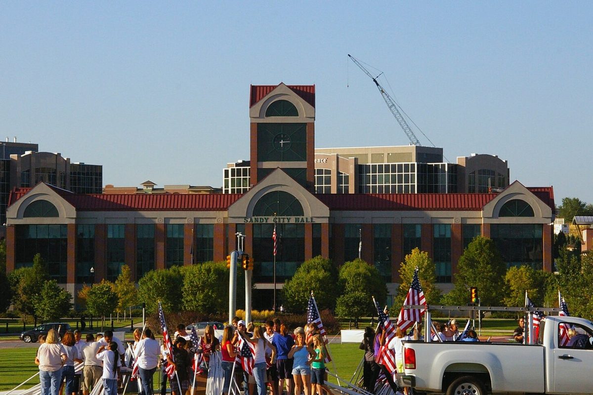 Sandy City Hall, a brick building with a red-roofed clock tower, as volunteers set up rows of American flags on the lawn and a white pickup waits at the curb