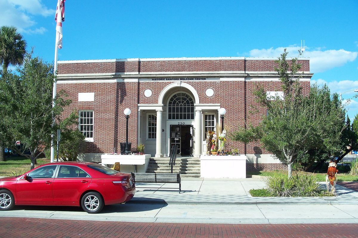Sanford's red brick Welcome Center, a former 1917 post office with an arched entrance, flag and lamps, with a red sedan parked on the brick street
