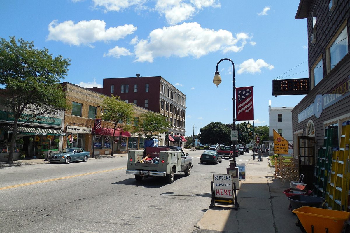 Main Street in Sanford on a sunny day, an old green pickup with a loaded bed driving past brick storefronts, with a flag banner on a lamppost and a sidewalk sign