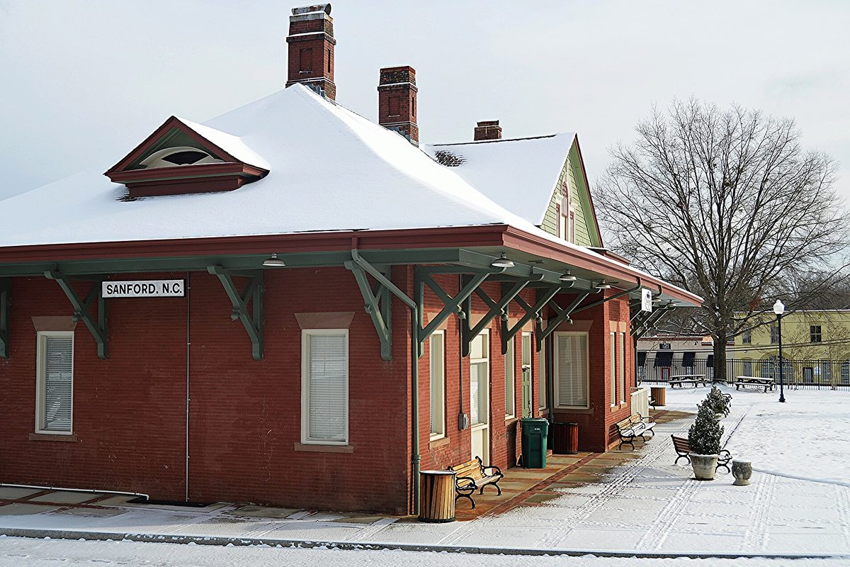 The red brick Sanford railroad depot, its SANFORD, N.C. sign over the platform, with snow on the roof and benches in Depot Park