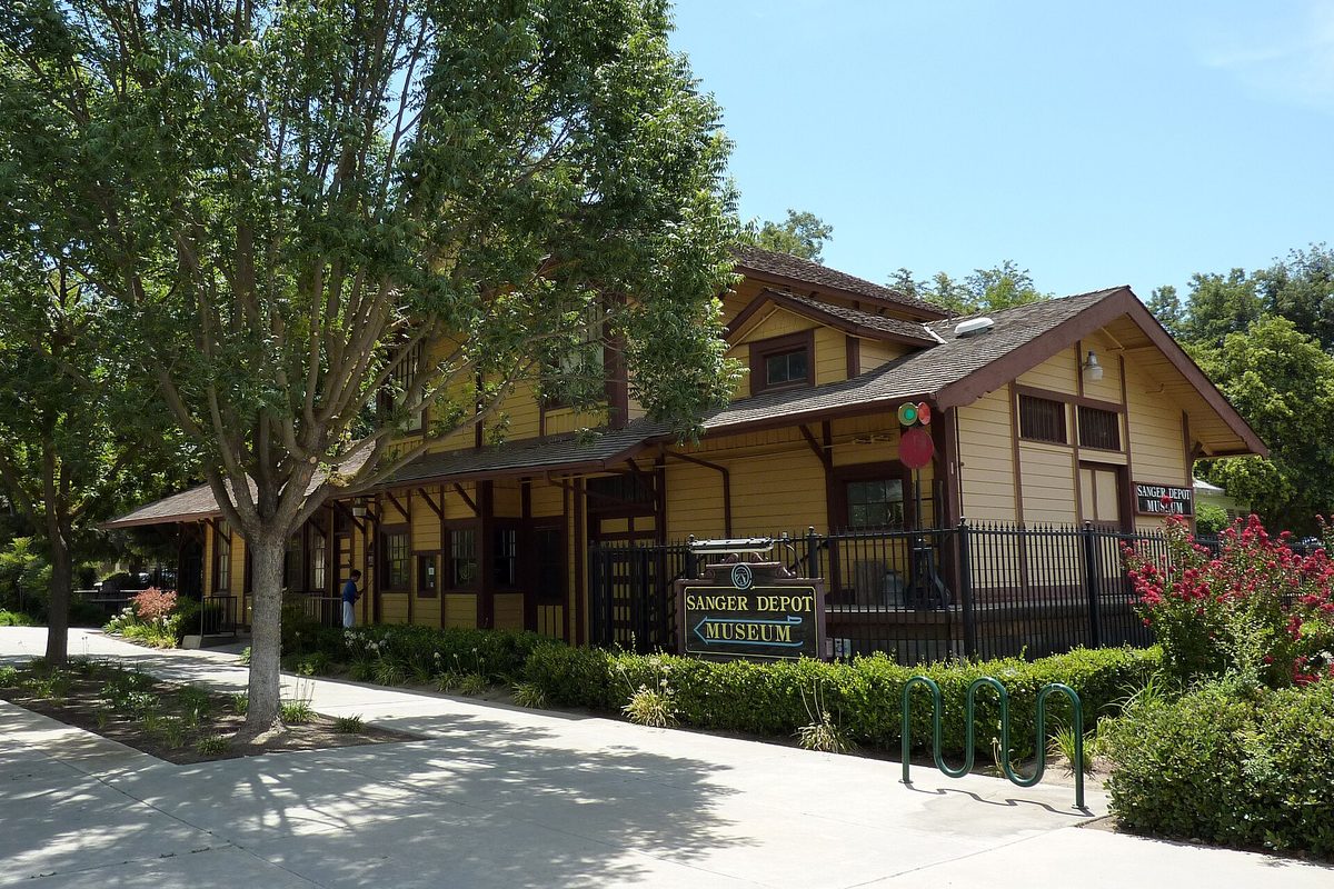The Sanger Depot Museum, a yellow wooden railroad depot with brown trim behind shade trees, a museum sign and a bike rack along the sidewalk