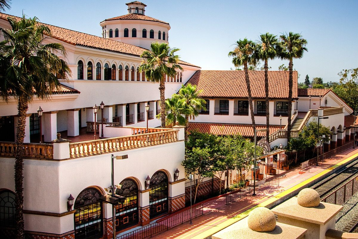 The white Spanish-style Santa Ana train station with red tile roofs, a round tower, palm trees and the platform beside the tracks