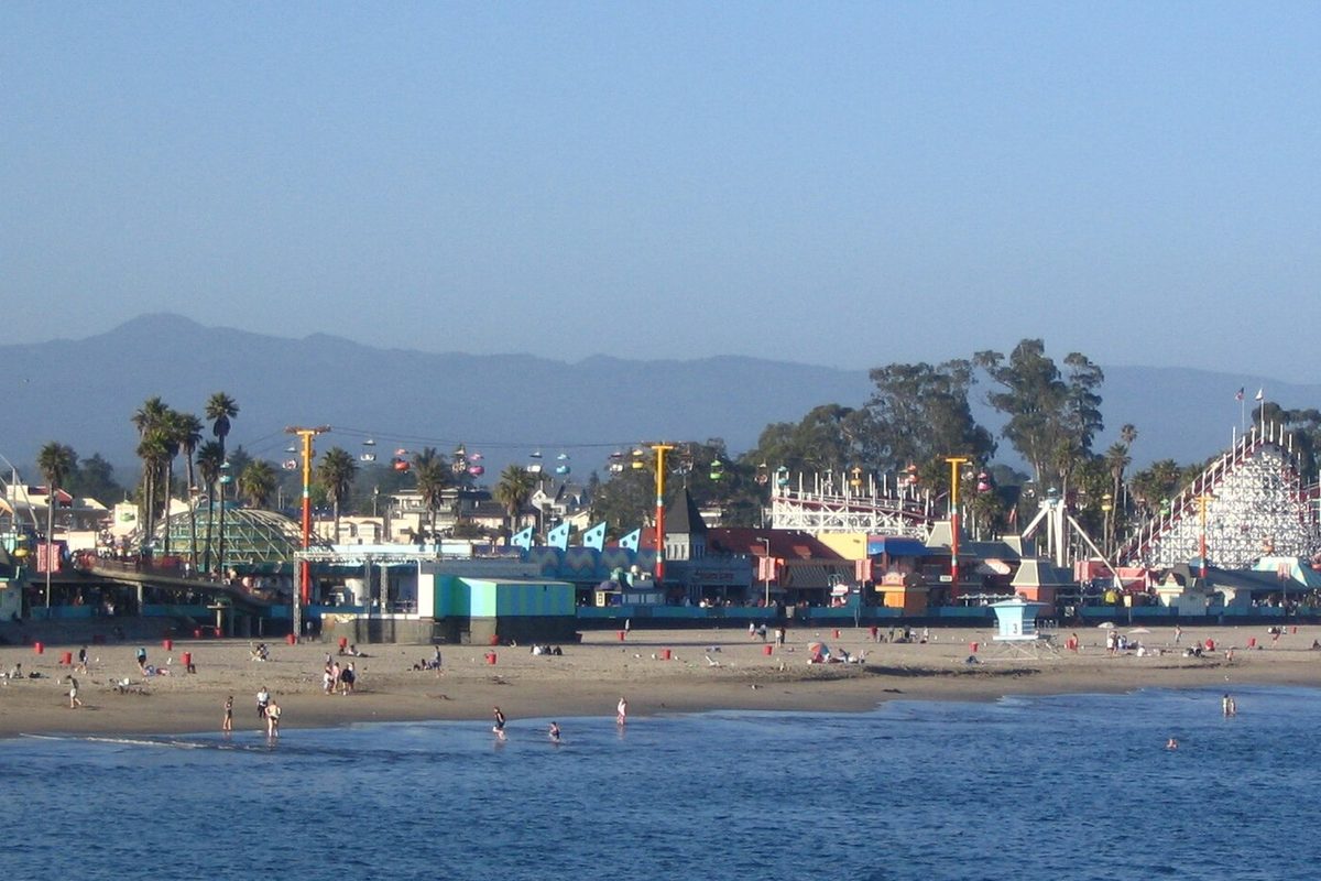The Santa Cruz Beach Boardwalk with its wooden roller coaster and rides behind a sandy beach and calm water, with hazy mountains beyond