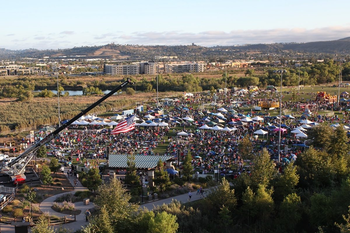 Crowds and white tents filling a park field at the Santee Salutes festival, seen from above, with an American flag hung from a crane and hills on the horizon