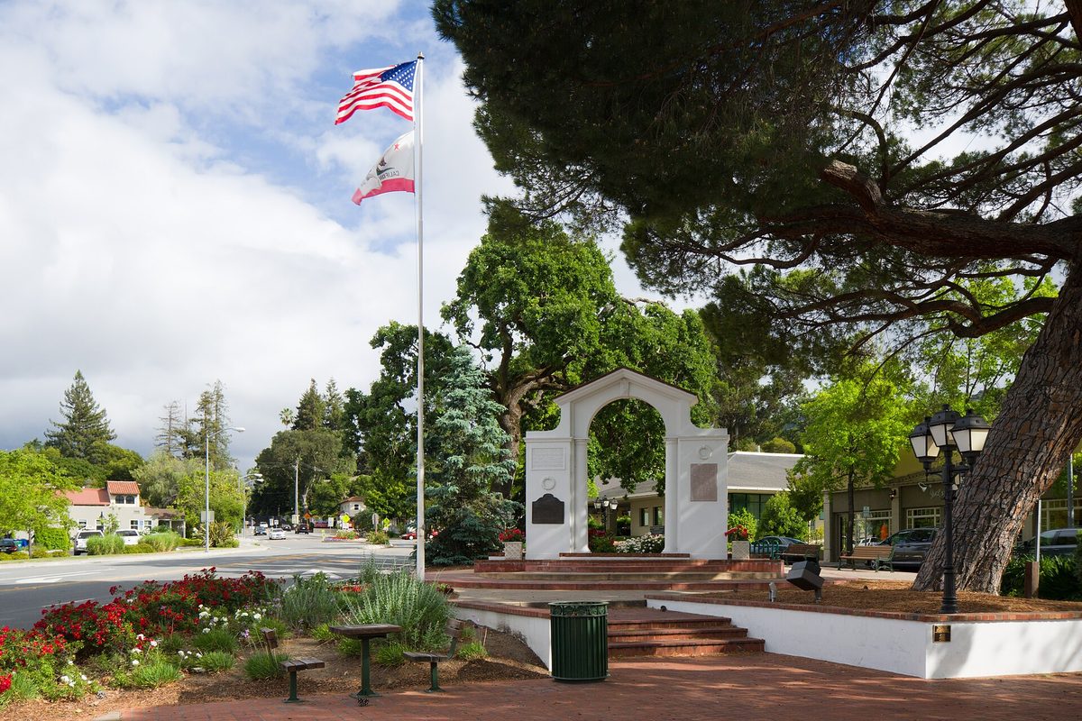Saratoga's white memorial arch in a brick plaza with red flowers, a flagpole flying the US and California flags, and tall pines and oaks behind