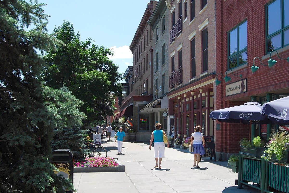 People walking a wide sidewalk past red brick storefronts, flower planters and café umbrellas in downtown Saratoga Springs