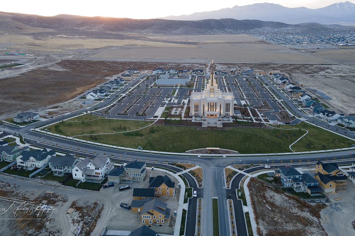 Aerial view at dusk of the Saratoga Springs Utah Temple with its white spire, ringed by new streets and houses, with open ground and mountains beyond
