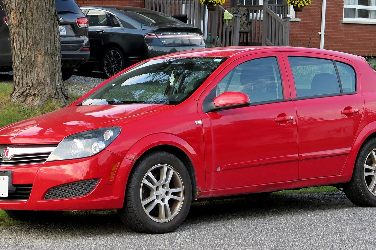 A red Saturn Astra four-door hatchback with alloy wheels, parked at a curb beside a tree trunk in front of houses