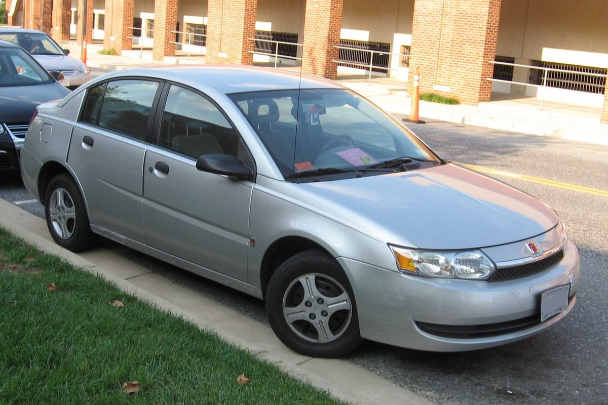 A silver 2003-2004 Saturn Ion sedan parked at a curb beside a strip of grass, a pink slip of paper on its dashboard