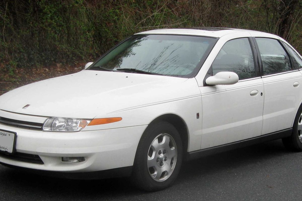 A white 2000-2002 Saturn L-Series sedan parked on a road beside a wooded slope
