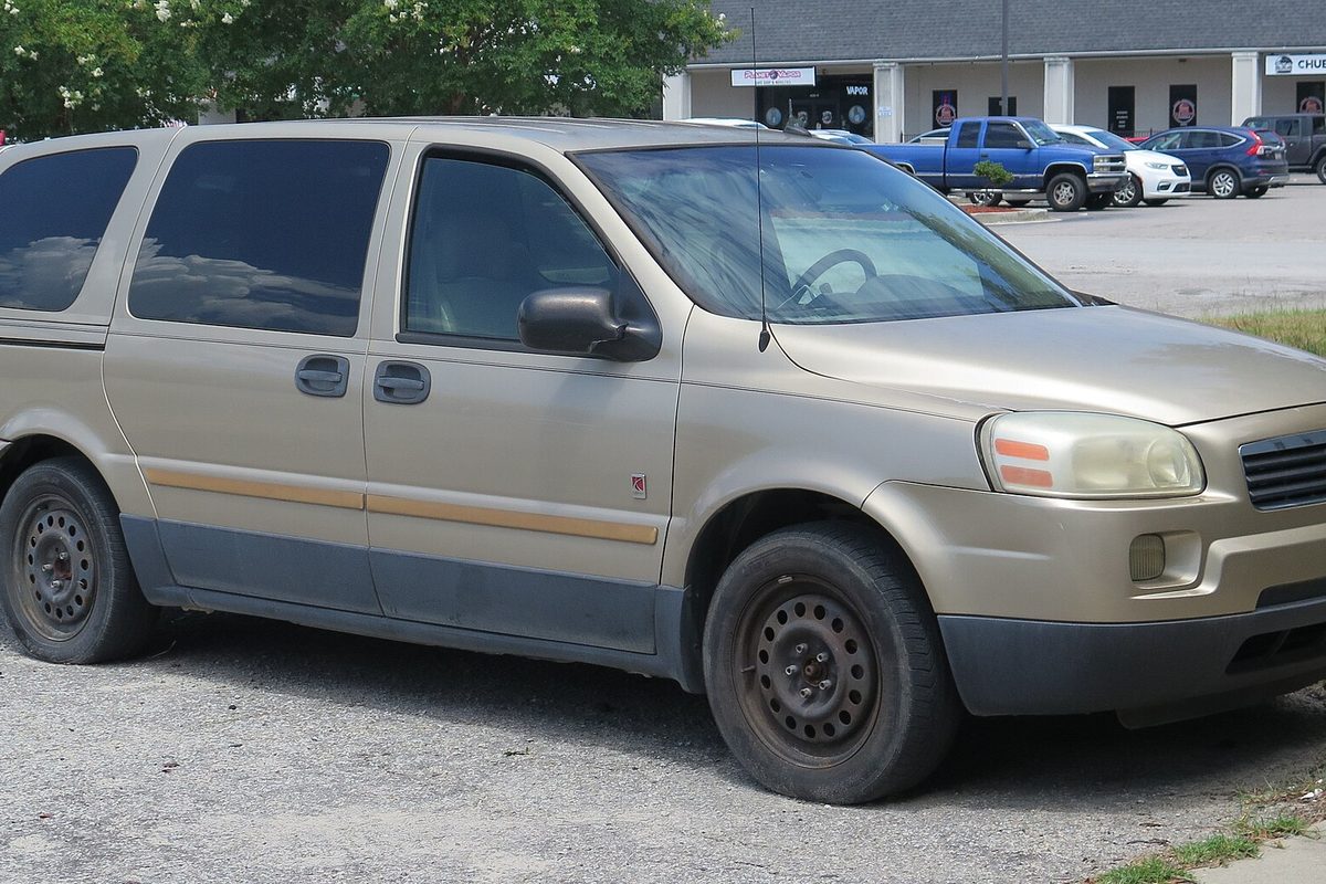 A tan 2005 Saturn Relay minivan with black steel wheels and gray lower trim, parked on gravel at a strip-mall lot