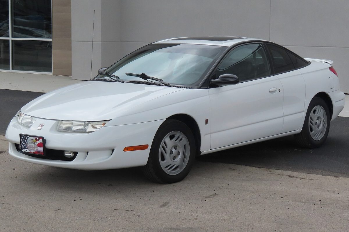 A white 1999 Saturn SC2 three-door coupe with silver wheel covers, parked on pavement in front of a building