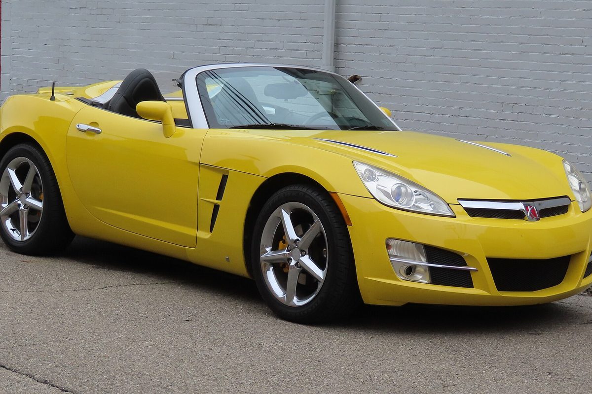 A yellow Saturn Sky two-seat convertible with the top down and chrome five-spoke wheels, parked beside a painted brick wall