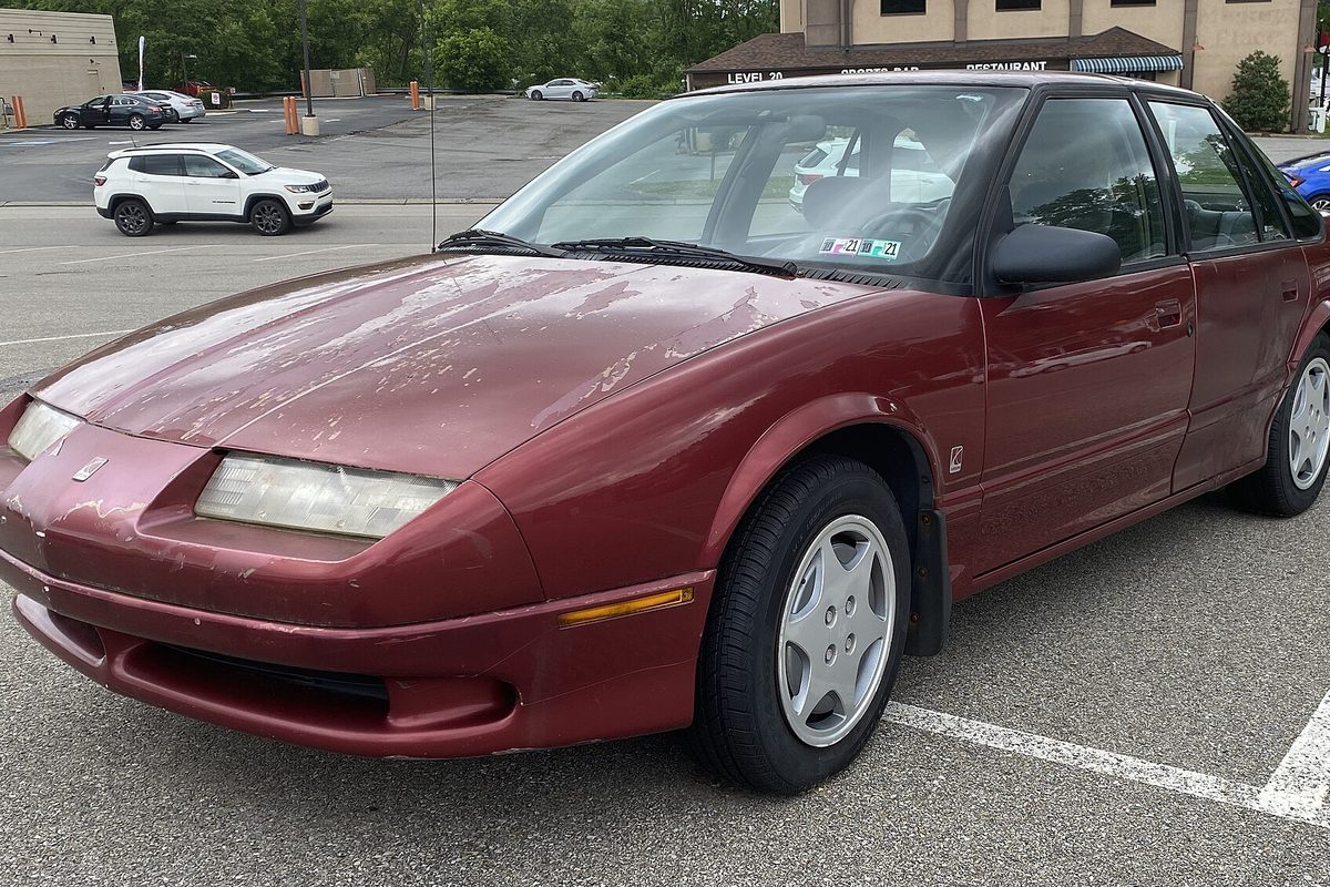 A red 1994 Saturn SL2 sedan with the clear coat peeling off its hood, parked in a lot