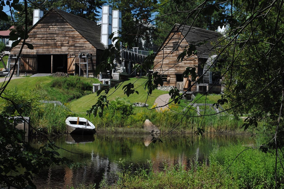 Weathered wooden mill buildings of the Saugus Iron Works on a grassy bank above the Saugus River, with a small white rowboat in the water