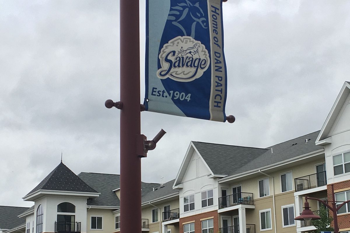 A blue street banner reading Savage, Home of Dan Patch, Est. 1904, on a lamppost in front of cream-sided apartment buildings under a gray sky
