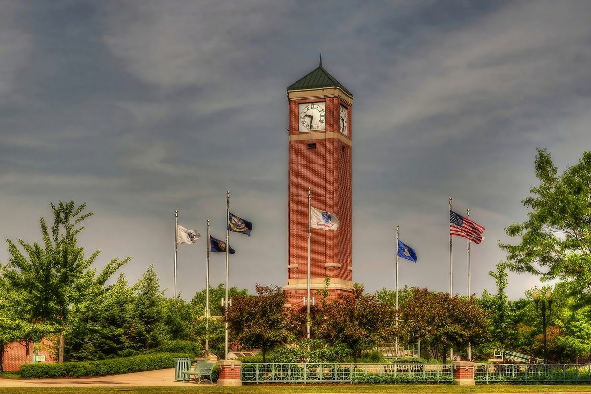 A red brick clock tower with a green pointed roof in Schaumburg, ringed by flagpoles flying the U.S. flag and other flags, with trees and a dark cloudy sky