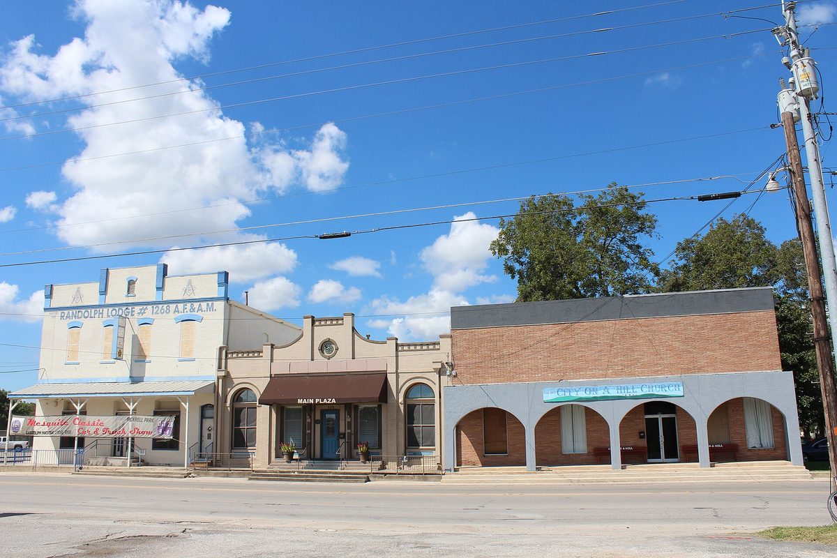 A row of old storefronts on Main Street in Schertz, including the Randolph Lodge building and an arched brick plaza front, under a blue sky with white clouds