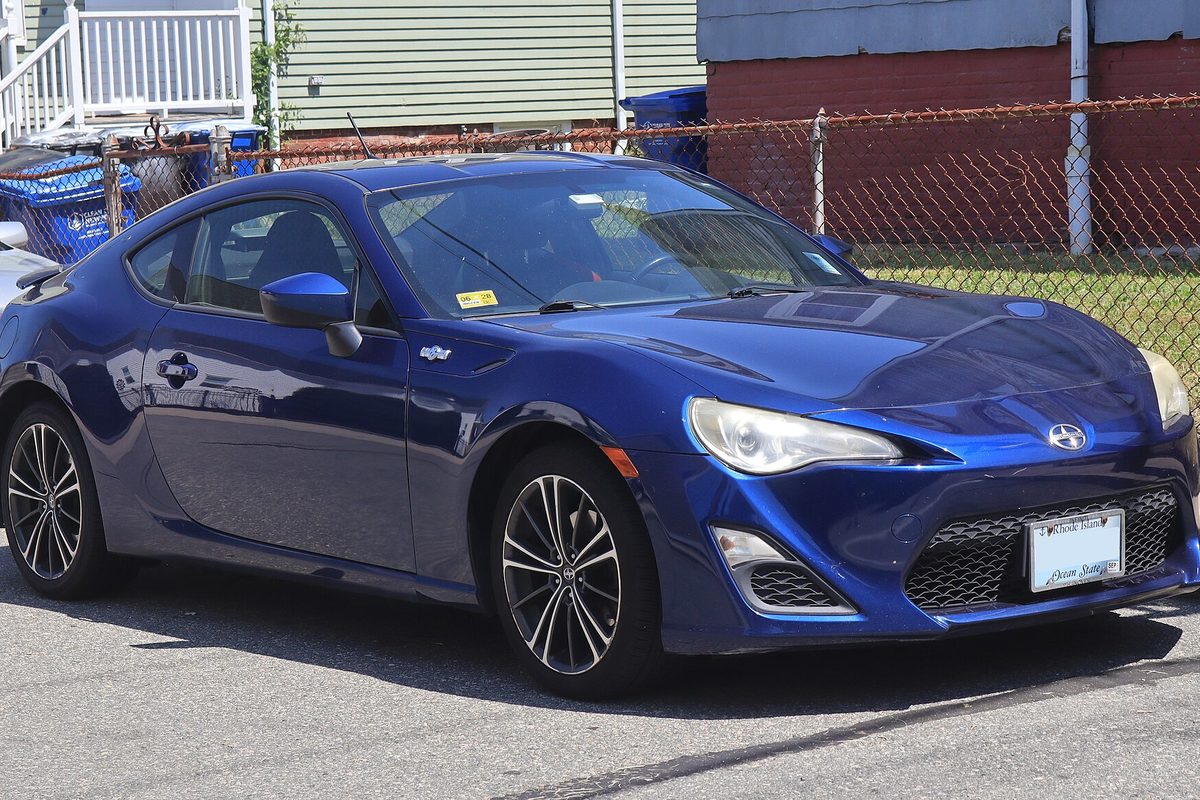 A dark blue 2013 Scion FR-S coupe with dark multi-spoke wheels parked beside a chain-link fence