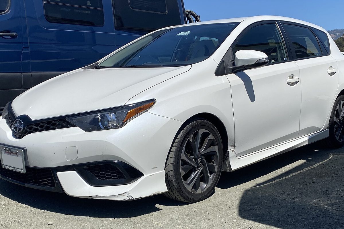 A white 2016 Scion iM hatchback seen from the front left, parked on gravel beside a blue van, with a scuffed lower front bumper