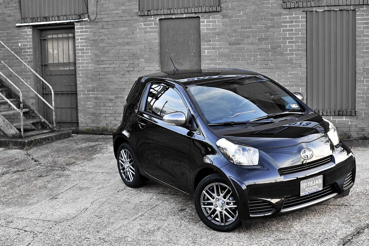 A black Scion iQ three-door minicar with silver alloy wheels parked in front of a brick building