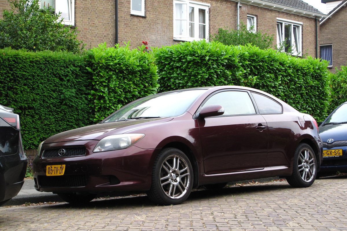 A dark red first-generation Scion tC coupe parked on a brick street in front of a tall green hedge