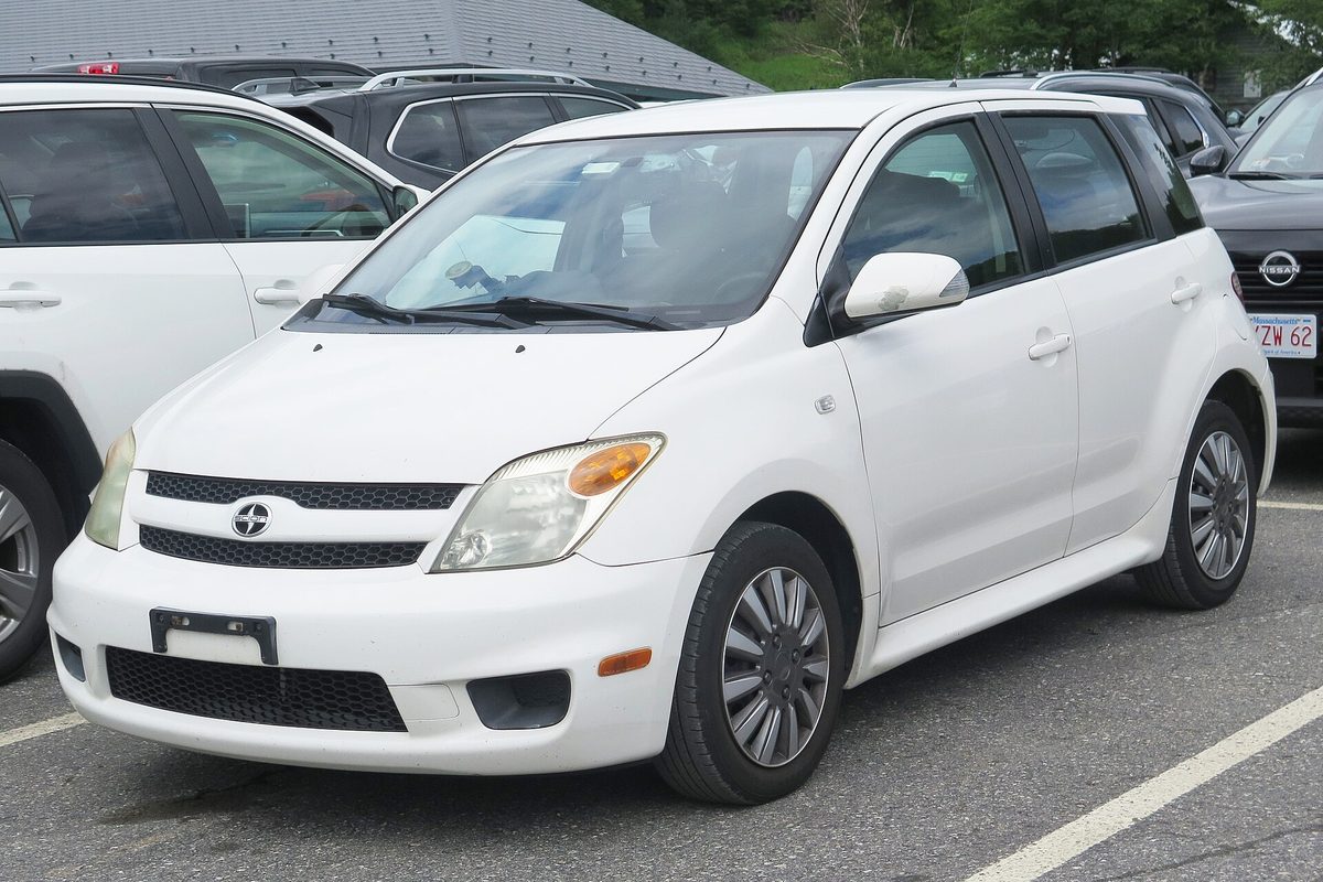 A white 2006 Scion xA five-door hatchback parked in a parking lot between two SUVs