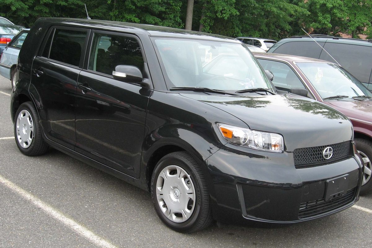 A black 2008 Scion xB, the boxy second-generation hatchback, parked in a lot next to a maroon car with trees behind it