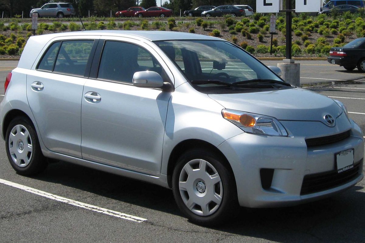 A silver 2008 Scion xD five-door hatchback with hubcaps parked in a lot, low shrubs behind it