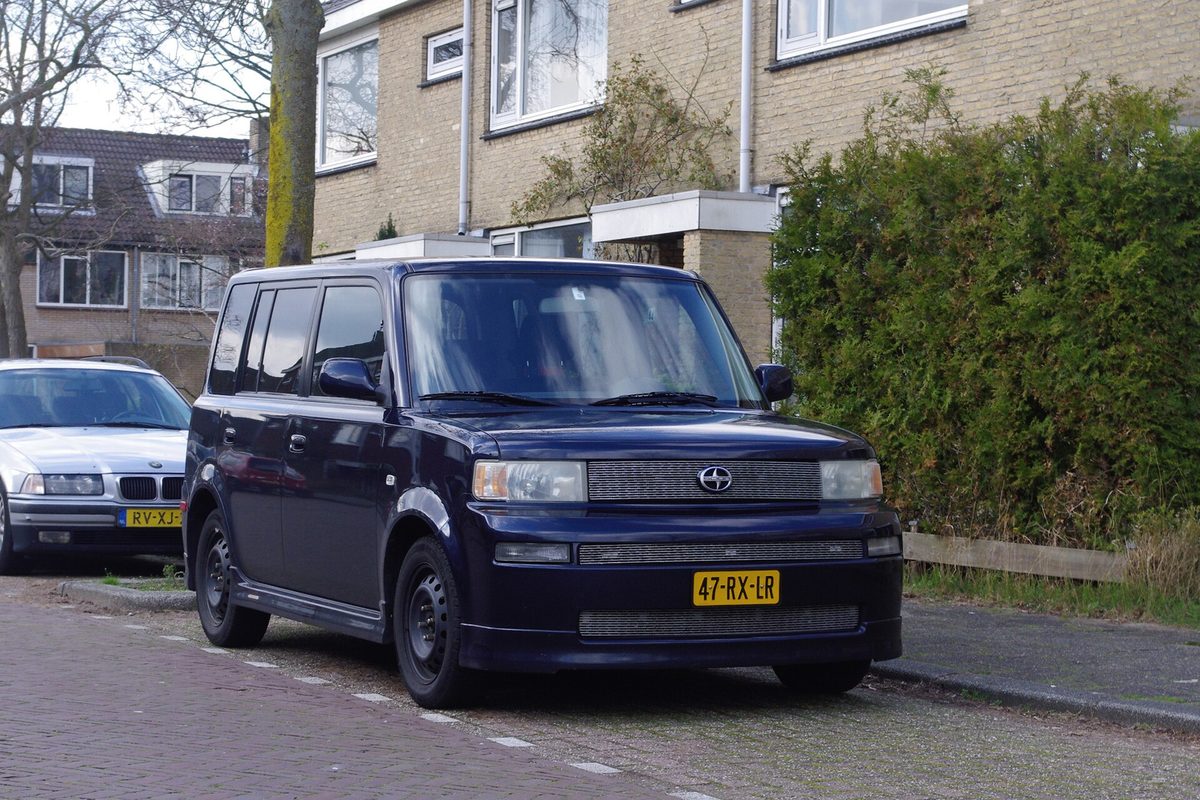 A dark blue first-generation Scion xB, the boxy compact wagon, parked at the curb of a brick street in front of row houses