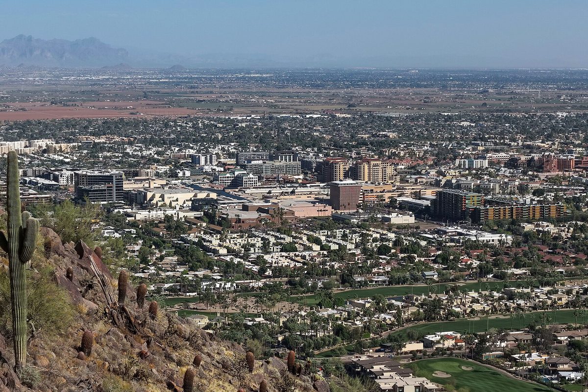 Downtown Scottsdale's mid-rise buildings and golf course greens seen from the rocky slope of Camelback Mountain, with a saguaro in the foreground and mountains on the horizon