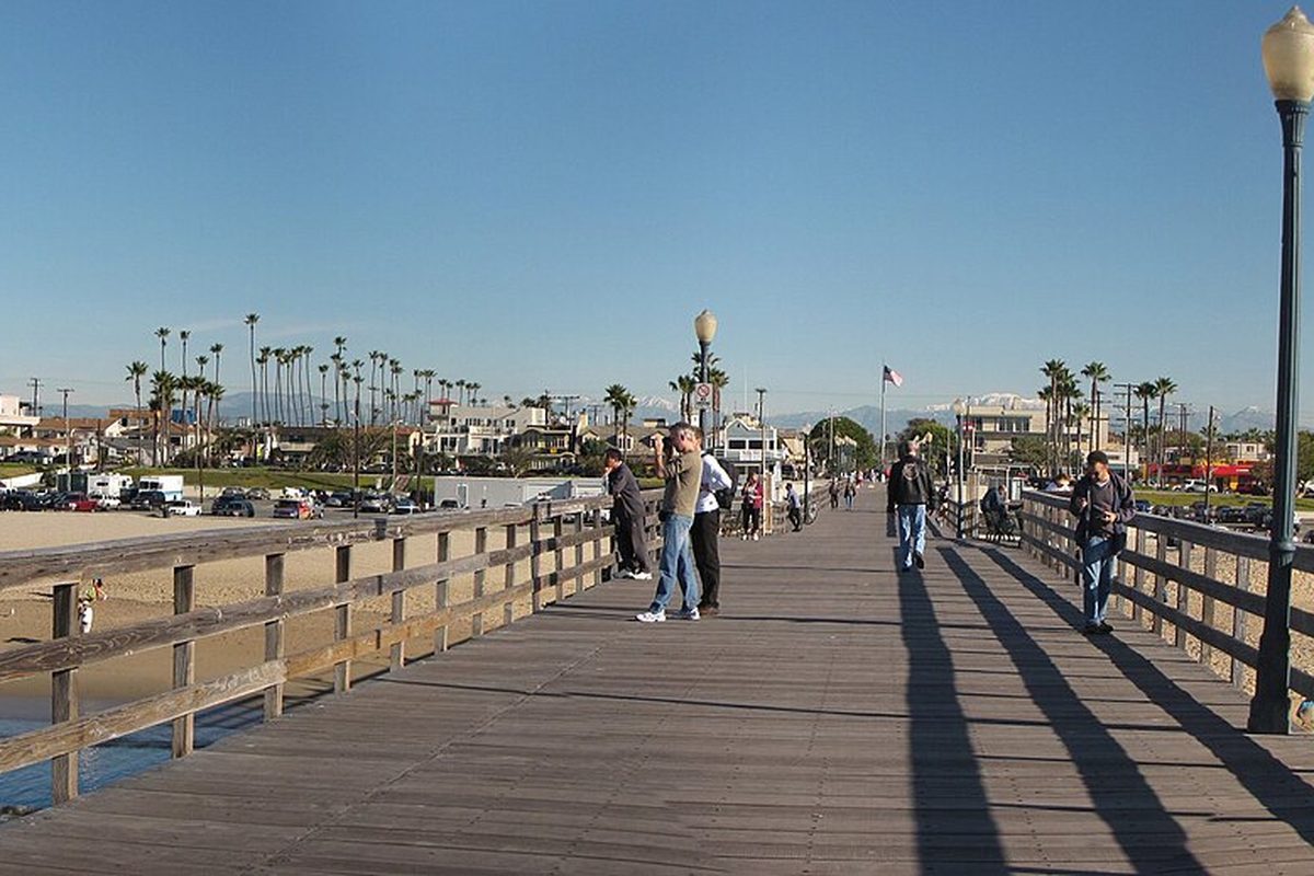 People walking along the wooden Seal Beach Pier on a clear day, with palm trees and beachfront homes along the shore
