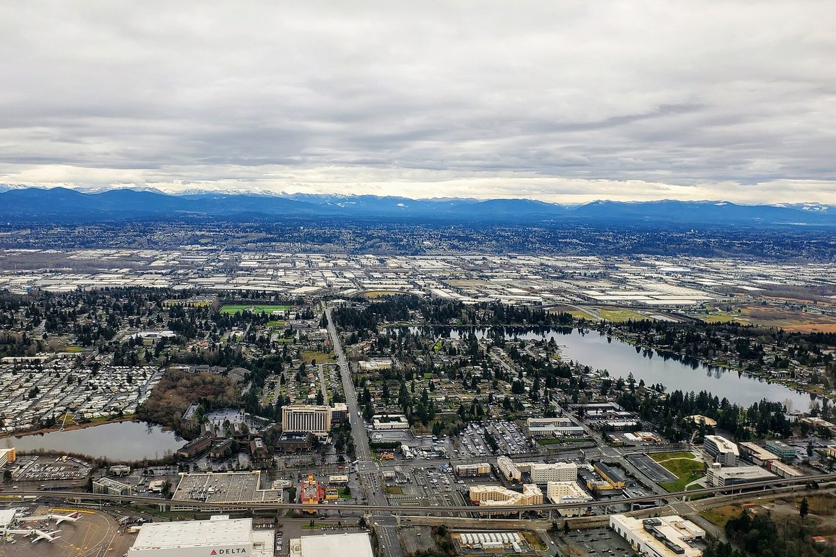 Aerial view over SeaTac with airliners and a hangar at the airport's edge, a highway running north between lakes and neighborhoods, and mountains under gray cloud