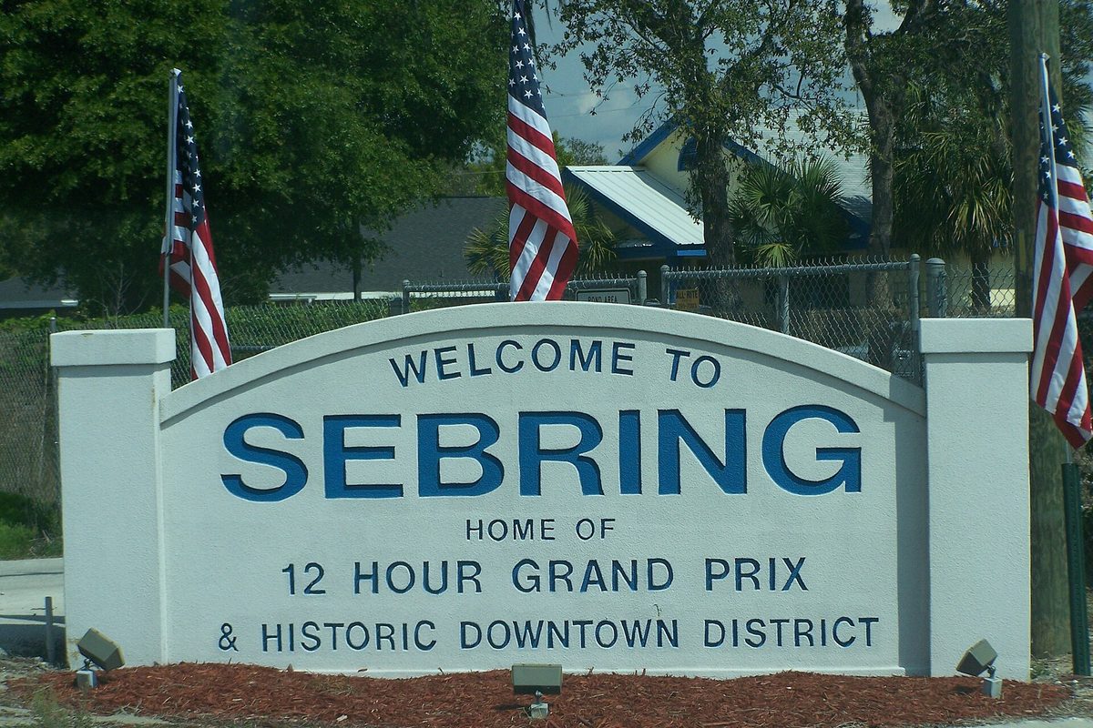 White Welcome to Sebring sign reading Home of 12 Hour Grand Prix and Historic Downtown District, flanked by American flags