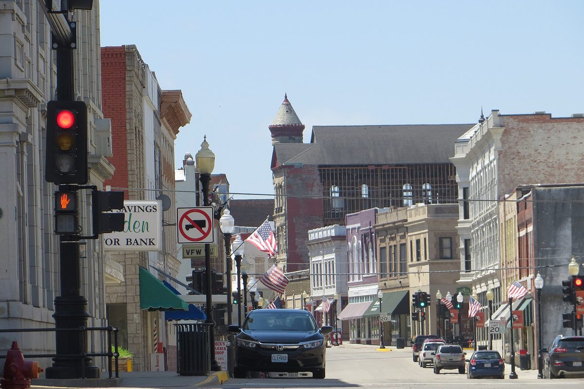 A downtown Sedalia street with a red traffic light in front, American flags on the lampposts, brick storefronts and a turreted building at the far end