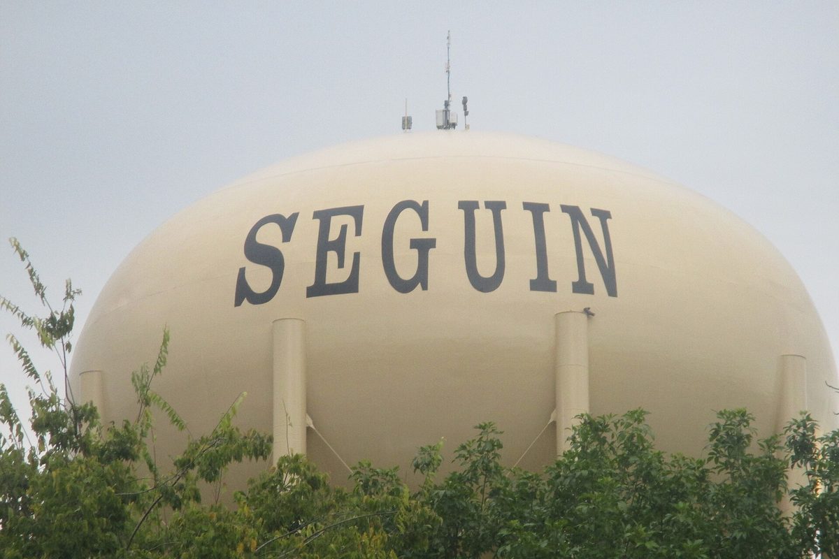 The pale yellow Seguin water tower, with SEGUIN painted in large dark letters, rising above green treetops under a hazy sky
