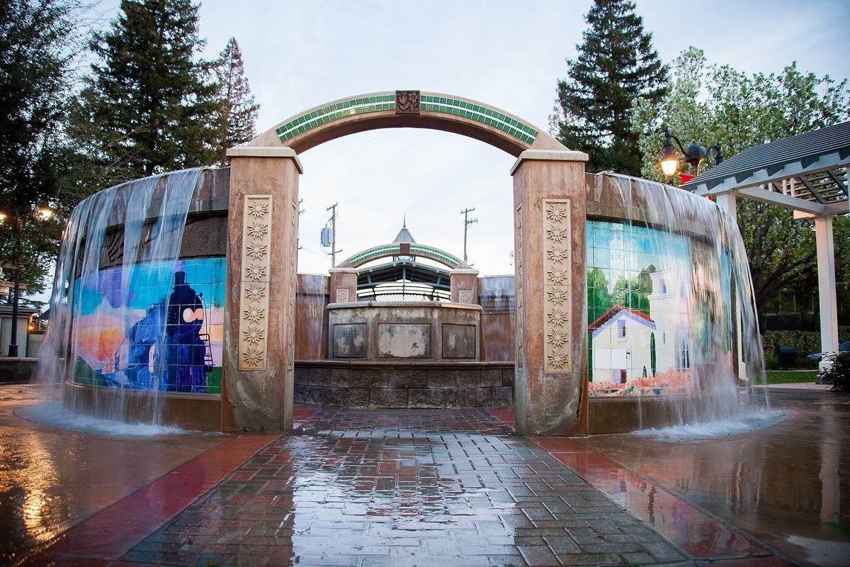 The fountain in downtown Shafter: water spilling over two tiled walls with murals of a locomotive and a white house, under a green arch, on a wet brick plaza